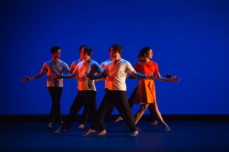 Members of Mark Morris Dance Group in bright orange costumes link at the elbows and walk across the stage